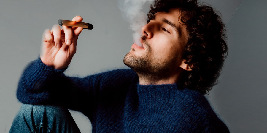 a man with curly hair wearing blue sweater and denim jeans vaping